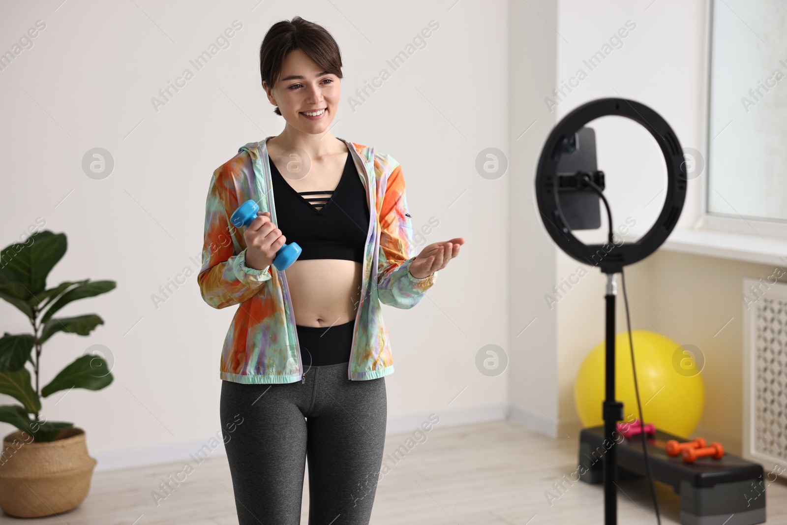 Photo of Happy sports blogger holding dumbbell while streaming online fitness lesson with smartphone at home