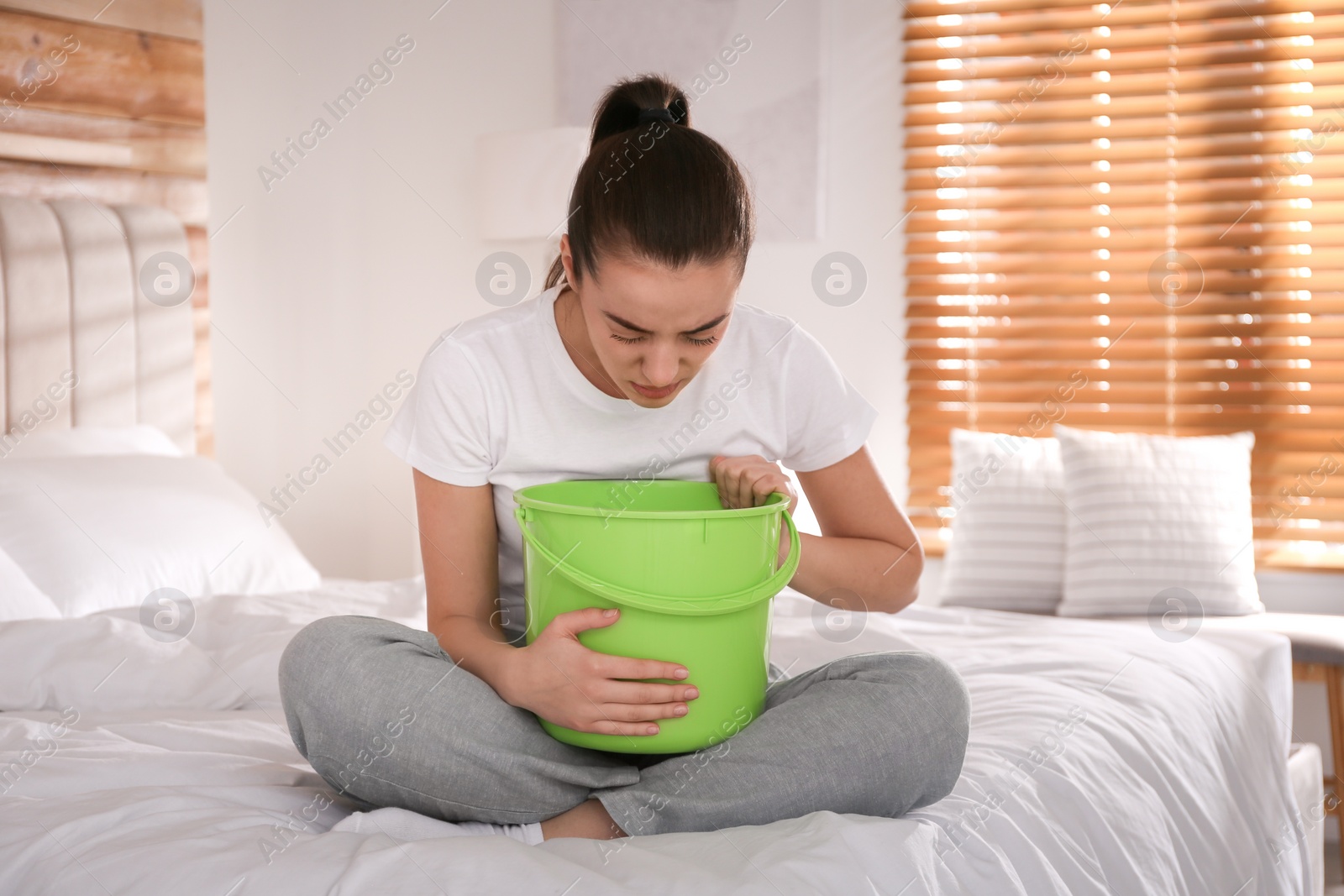 Photo of Young woman with bucket suffering from nausea on bed at home. Food poisoning