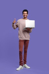 Smiling man with laptop showing thumb up on purple background