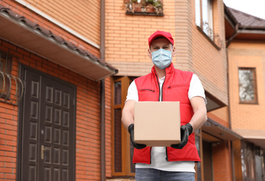 Courier in protective mask and gloves with box near house outdoors. Delivery service during coronavirus quarantine