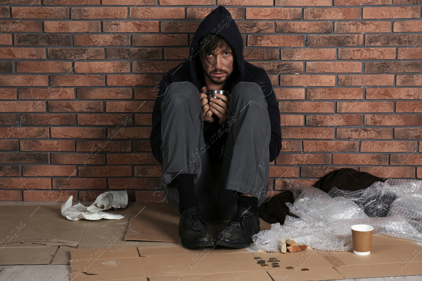 Photo of Poor man sitting with mug on floor near brick wall