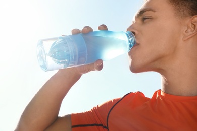 Young sporty man drinking water from bottle against blue sky on sunny day