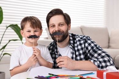 Photo of Dad and son covering mouth with paper mustaches at home