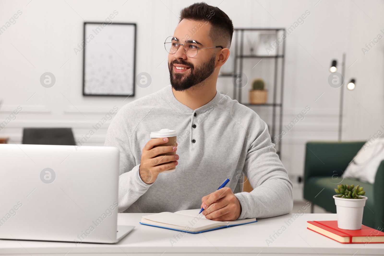 Photo of Young man with cup of coffee writing in notebook at white table indoors