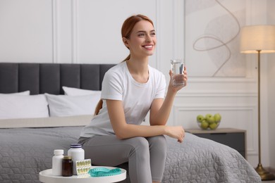 Happy young woman holding glass of water near table with pills and measuring tape in room. Weight loss