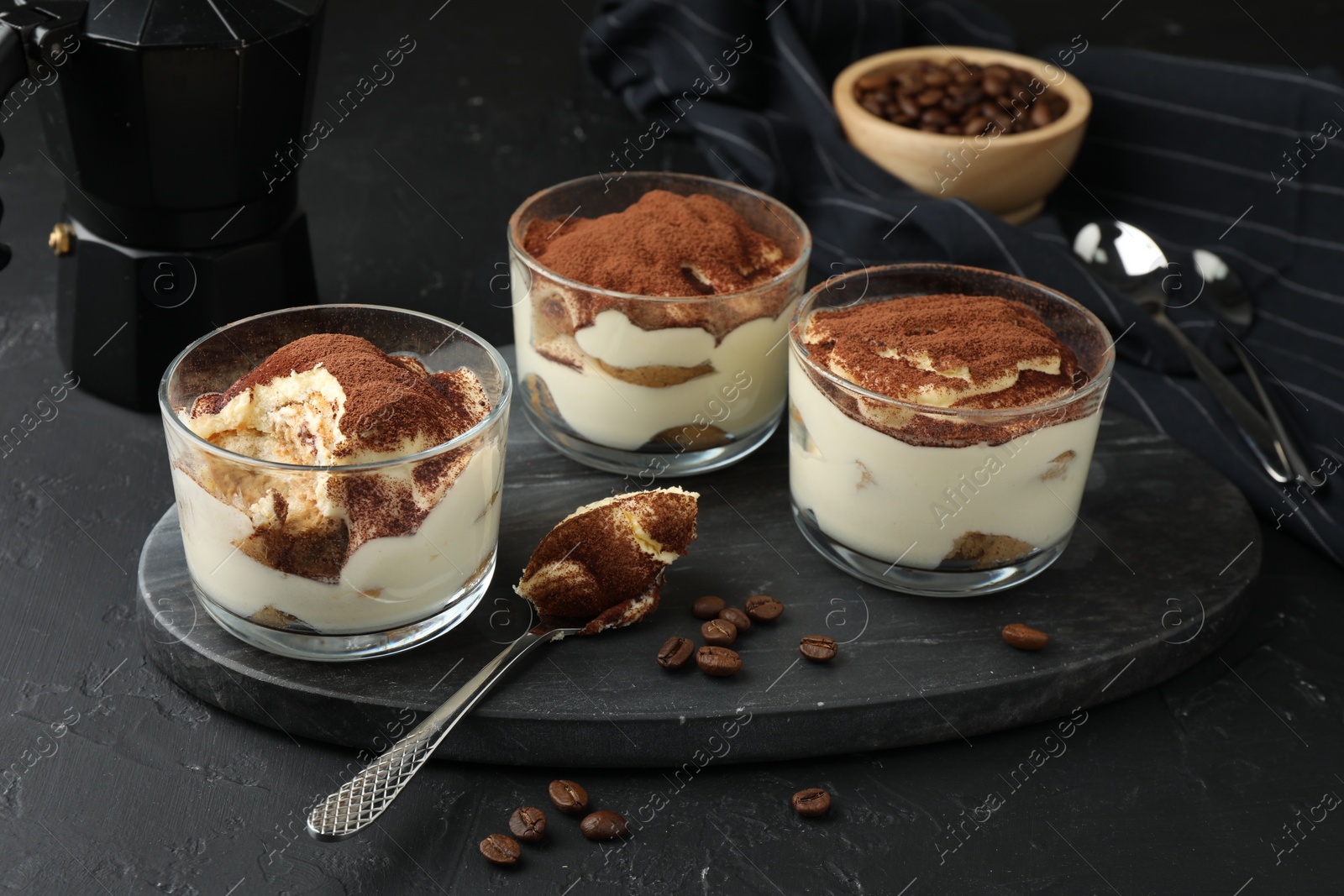 Photo of Delicious tiramisu in glasses, spoon and coffee beans on black table