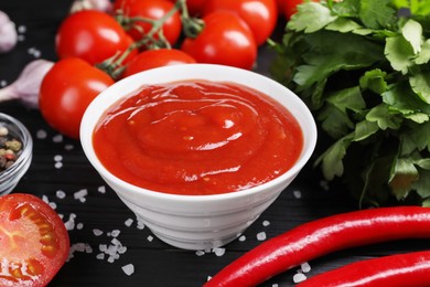 Photo of Delicious ketchup in bowl, products and salt on black wooden table, closeup. Tomato sauce