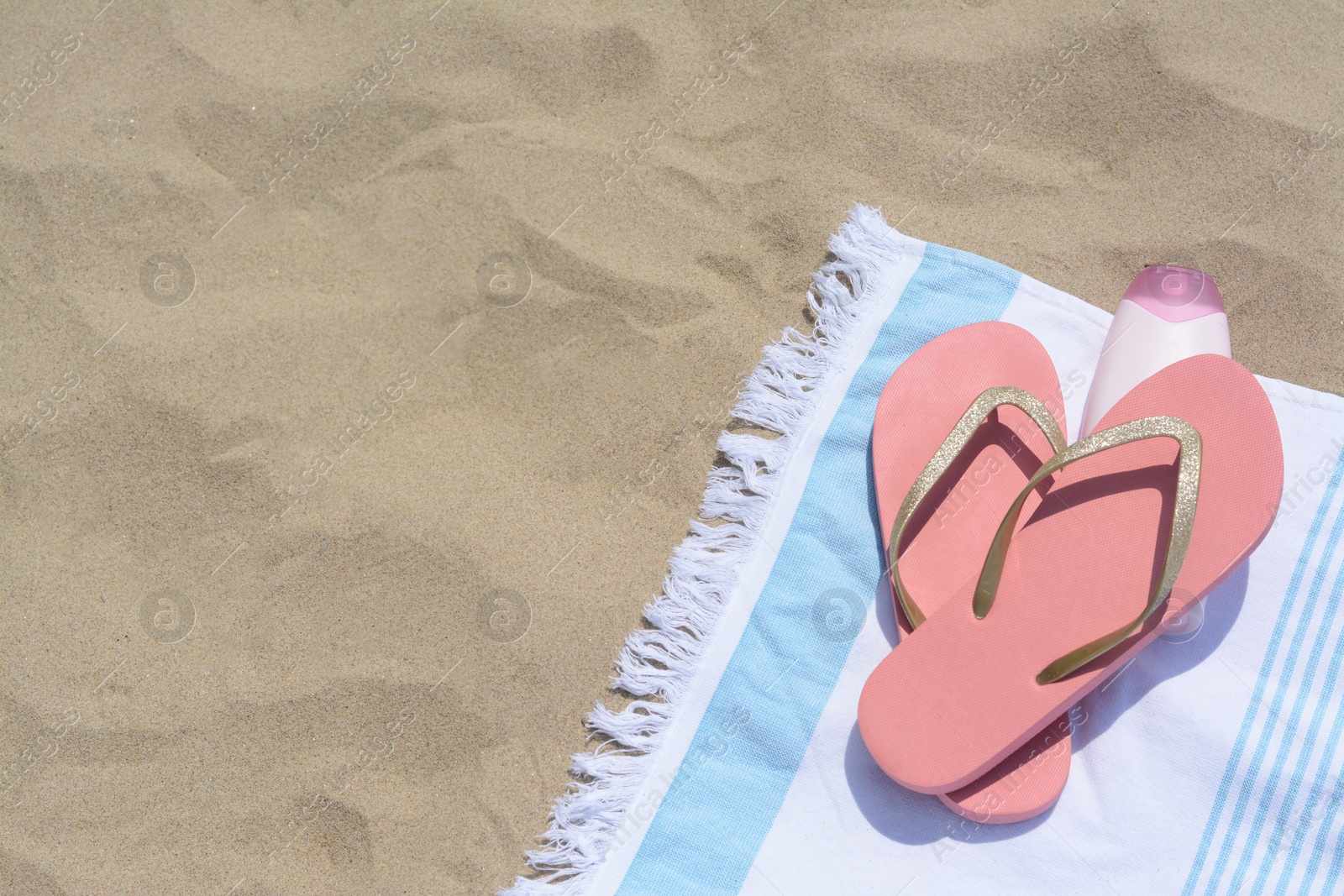 Photo of Striped towel with bottle of sunblock and flip flops on sandy beach, flat lay. Space for text
