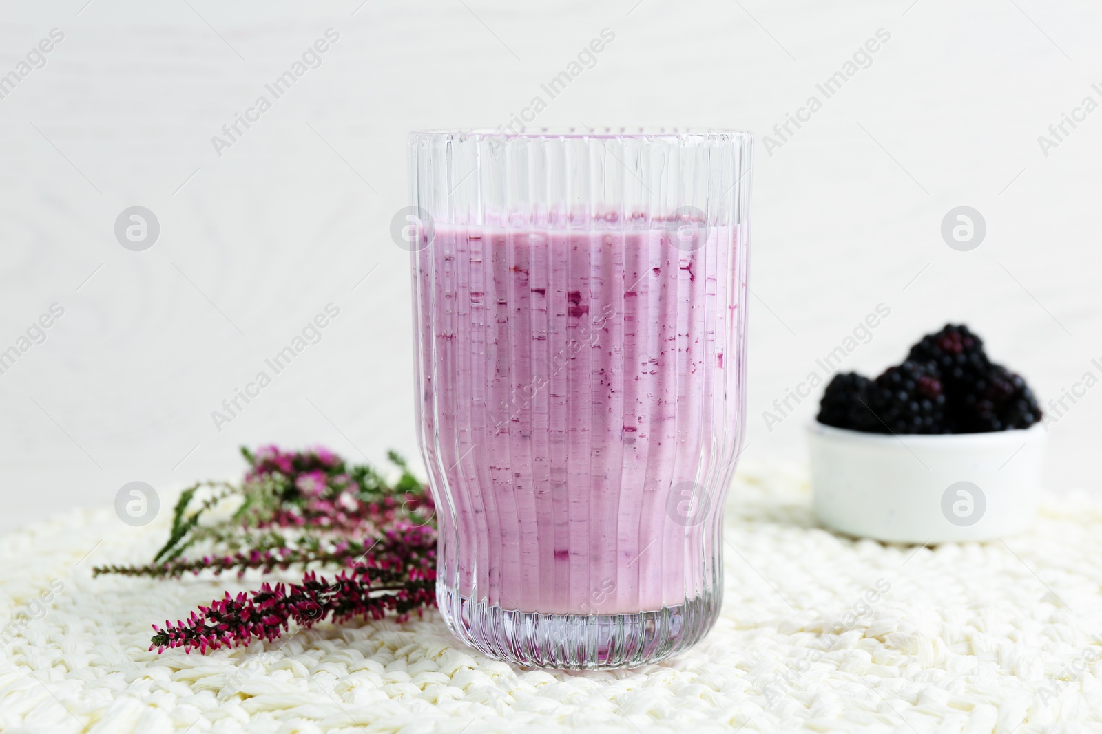 Photo of Glass of delicious blackberry smoothie, fresh berries and flowers on white wooden background