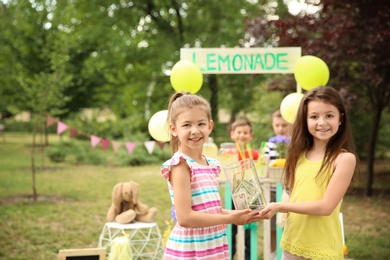 Photo of Little girls holding jar with money near lemonade stand in park