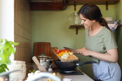 Photo of Young woman putting cut vegetables into frying pan in kitchen