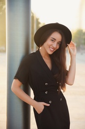 Beautiful young woman in stylish black dress and hat on city street