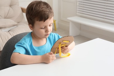 Photo of Little boy tying shoe lace using training cardboard template at white table indoors