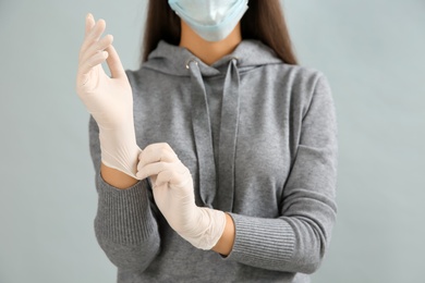 Woman in protective face mask putting on medical gloves against grey background, closeup