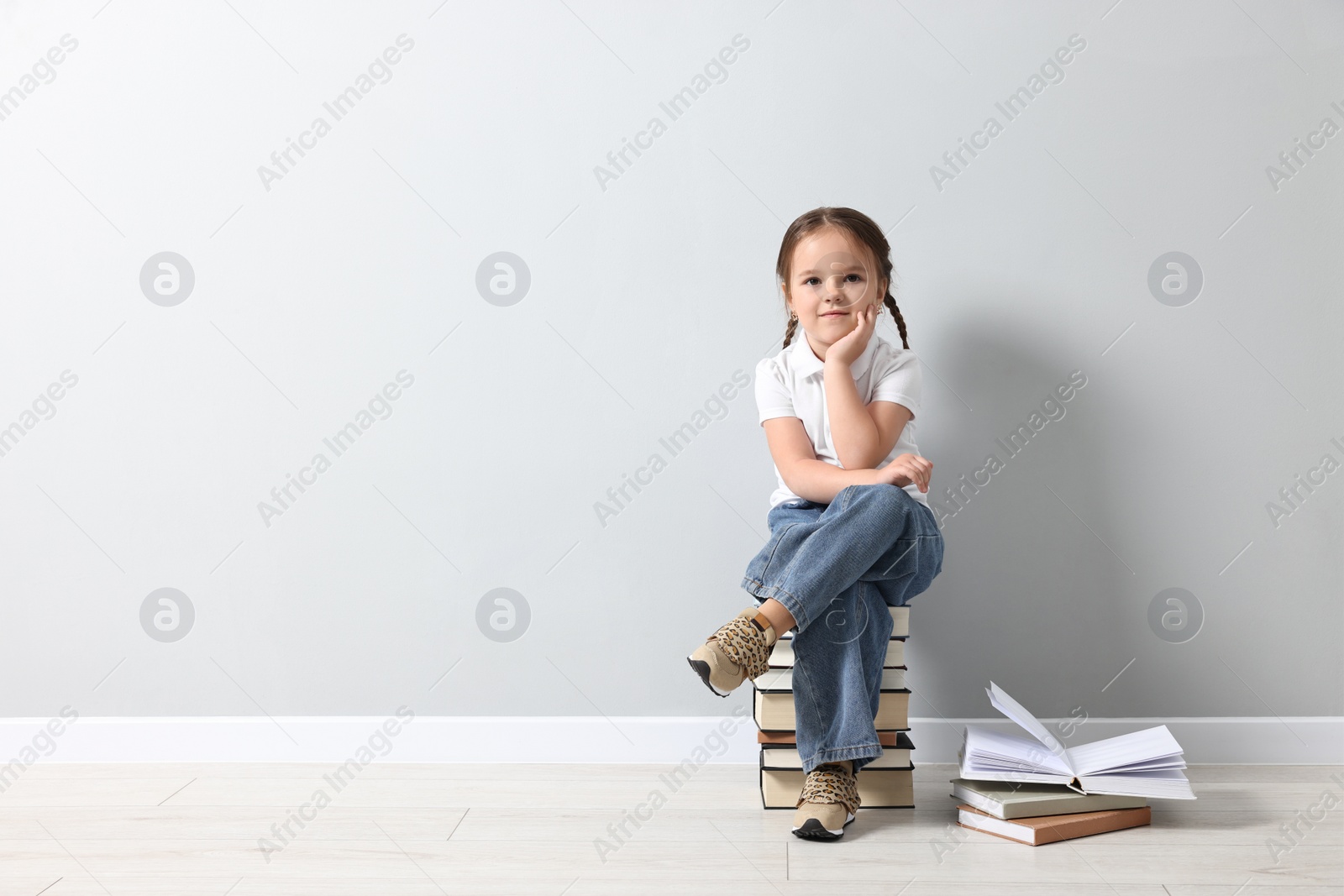 Photo of Cute little girl sitting on stack of books near light grey wall. Space for text