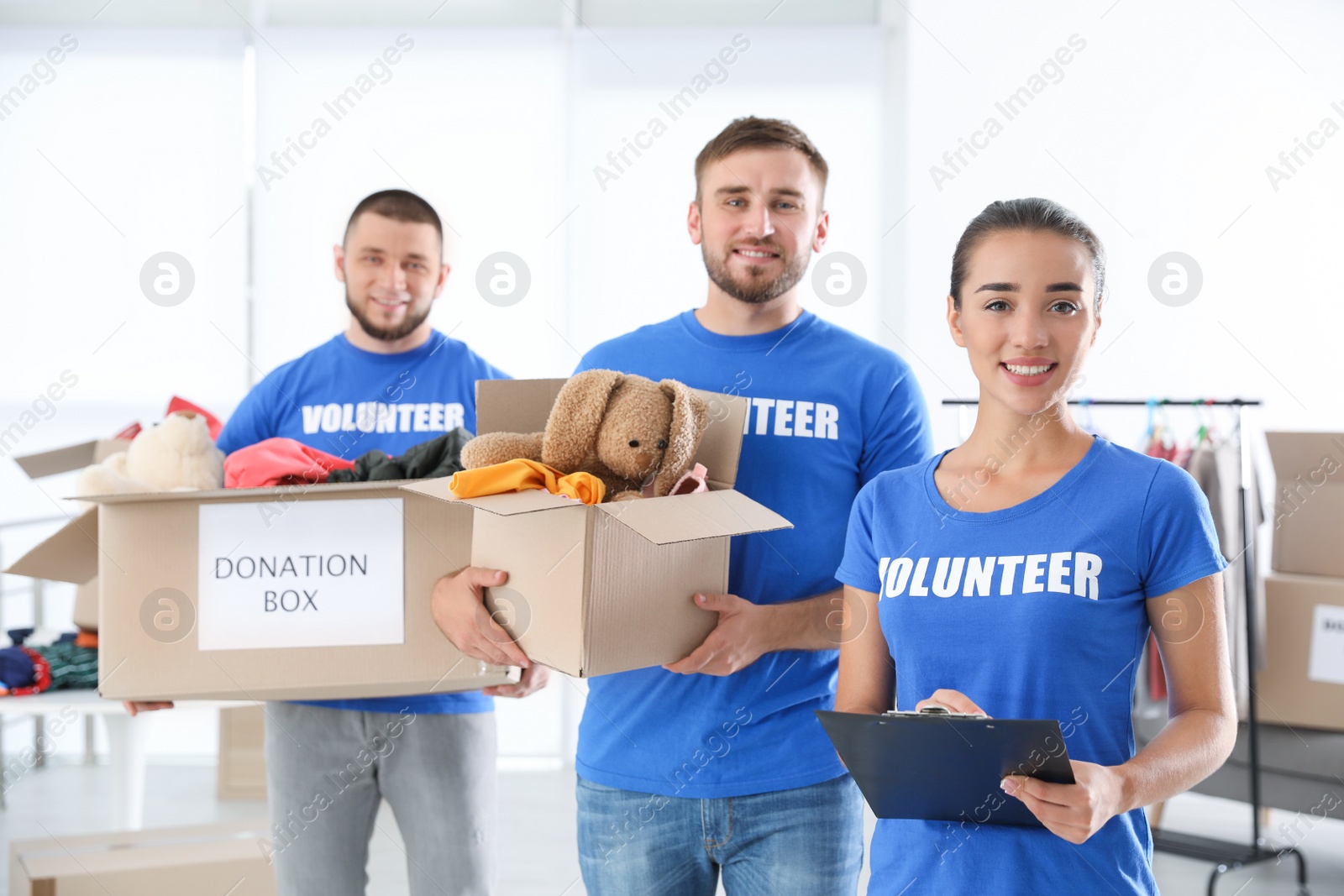 Photo of Young female volunteer with clipboard and her team holding boxes full of donations indoors