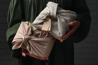 Photo of Furoshiki technique. Woman holding gifts wrapped in fabric with thuja branches near dark brick wall, closeup