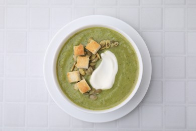 Delicious broccoli cream soup with croutons, sour cream and pumpkin seeds on white tiled table, top view