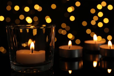 Photo of Glass holder with burning tea candle against blurred lights in darkness, closeup. Space for text