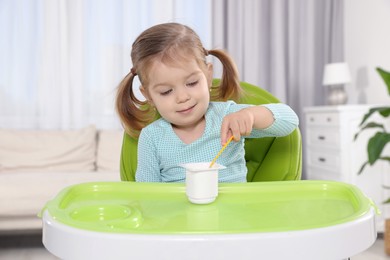 Cute little child eating tasty yogurt from plastic cup with spoon in high chair indoors