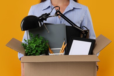 Photo of Unemployed woman holding box with personal office belongings on orange background, closeup