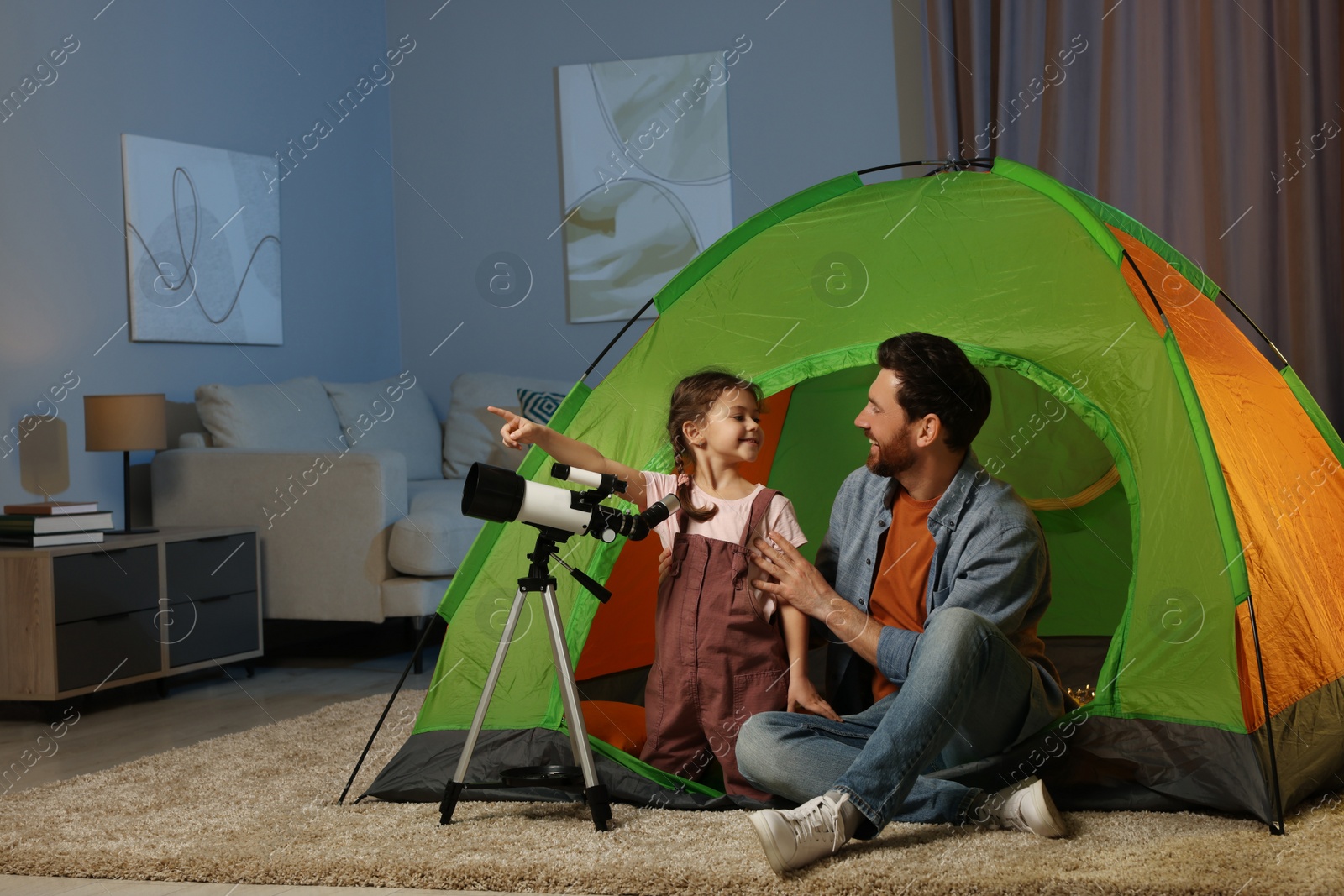 Photo of Father and his daughter using telescope to look at stars while sitting in camping tent indoors