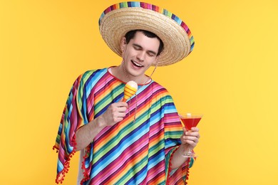 Photo of Young man in Mexican sombrero hat and poncho with cocktail and maracas on yellow background