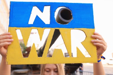 Photo of Woman holding poster in colors of Ukrainian flag with words No War near tank barrel on city street, closeup