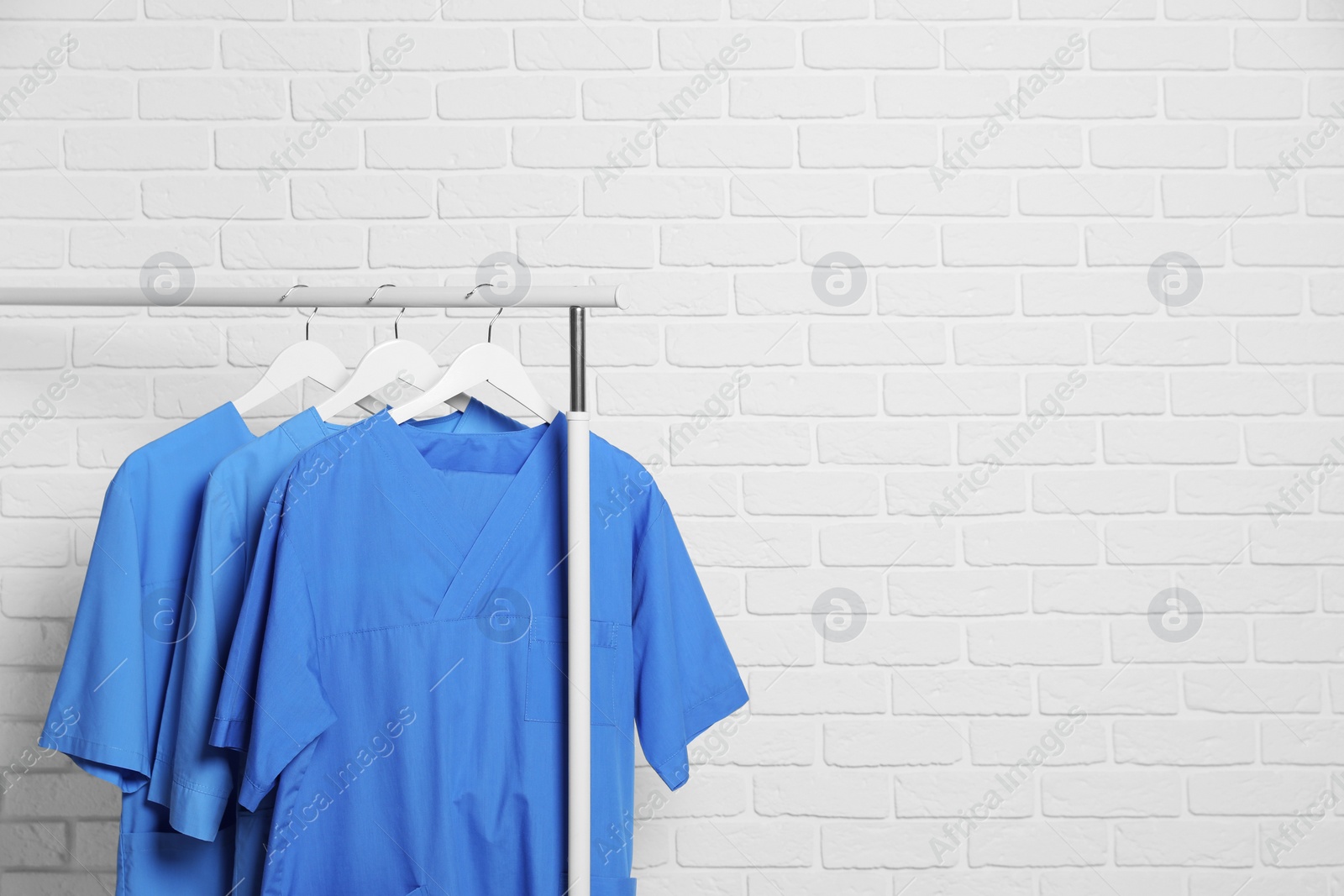 Photo of Medical uniforms hanging on rack near white brick wall. Space for text