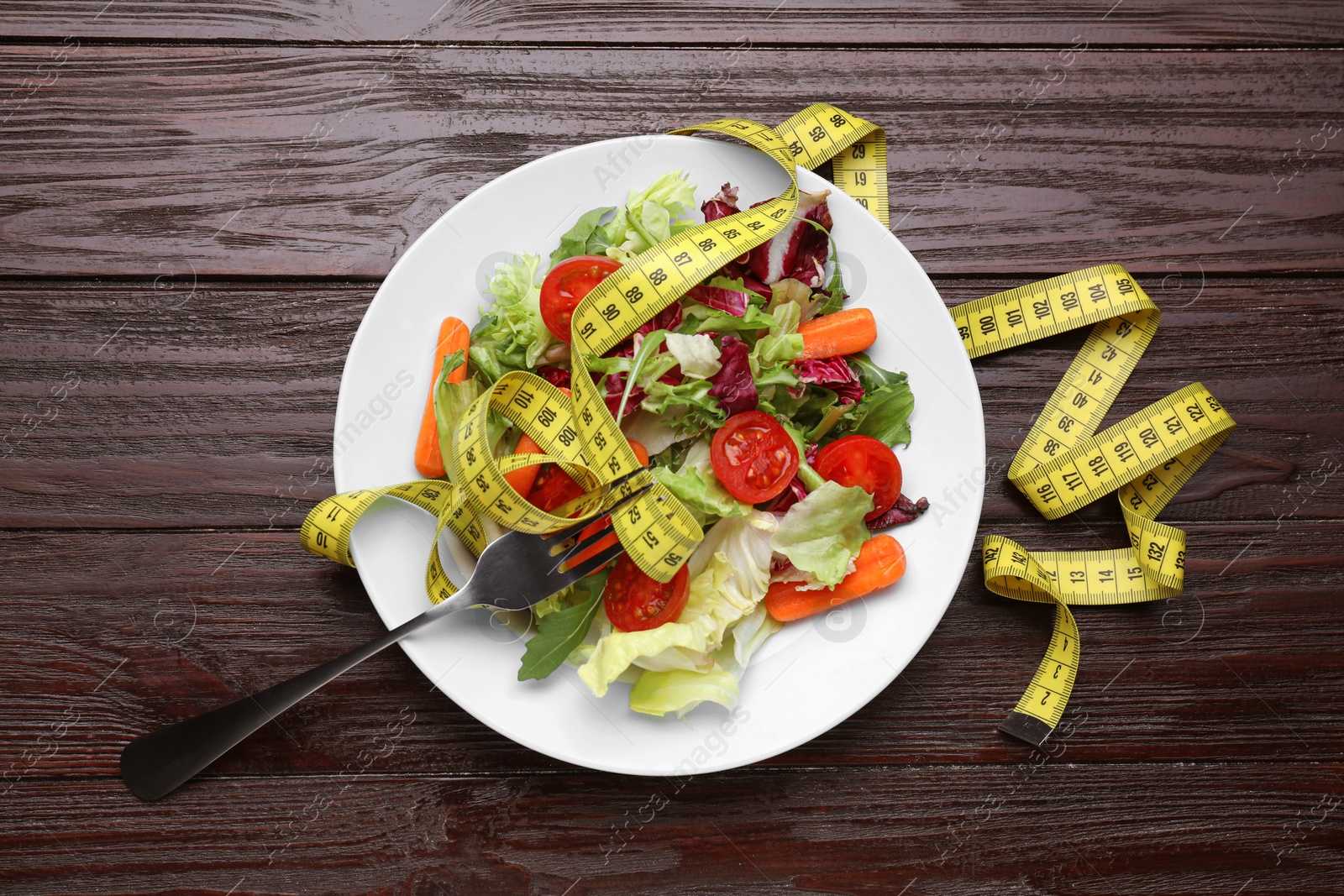 Photo of Plate with fresh vegetable salad, fork and measuring tape on wooden table, flat lay. Healthy diet concept