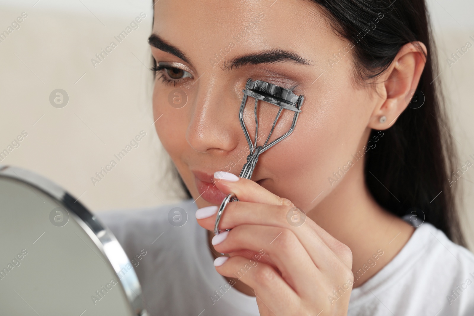 Photo of Beautiful young woman using eyelash curler in front of mirror indoors, closeup