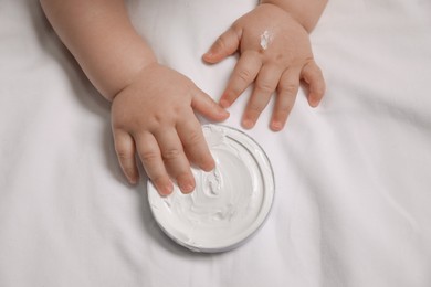 Photo of Cute little baby playing with jar of moisturizing cream on bed, closeup