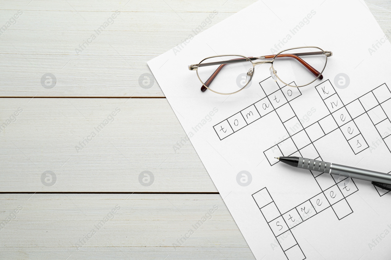 Photo of Crossword with answers, glasses and pen on white wooden table, top view. Space for text