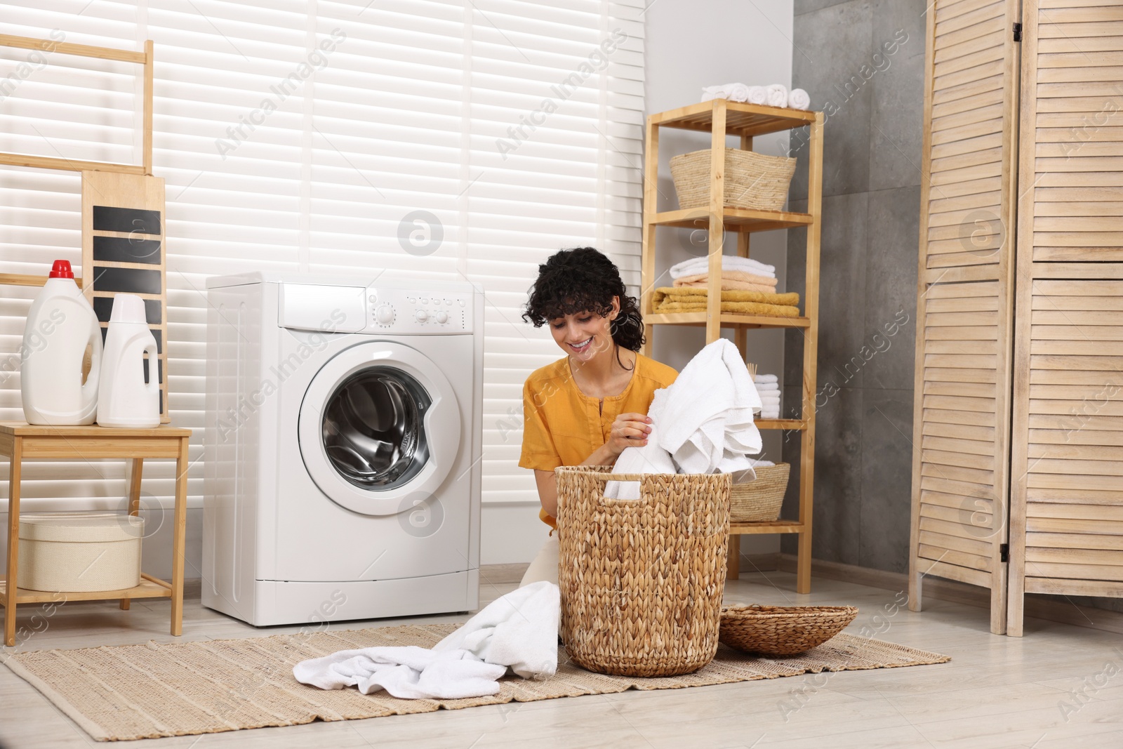 Photo of Happy woman taking laundry from basket near washing machine indoors