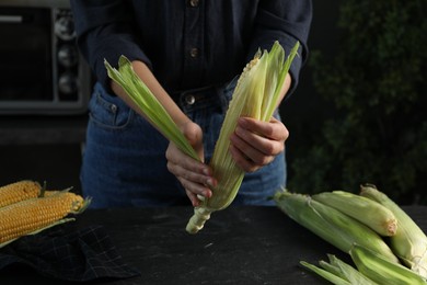 Photo of Woman husking corn cob at black table, closeup