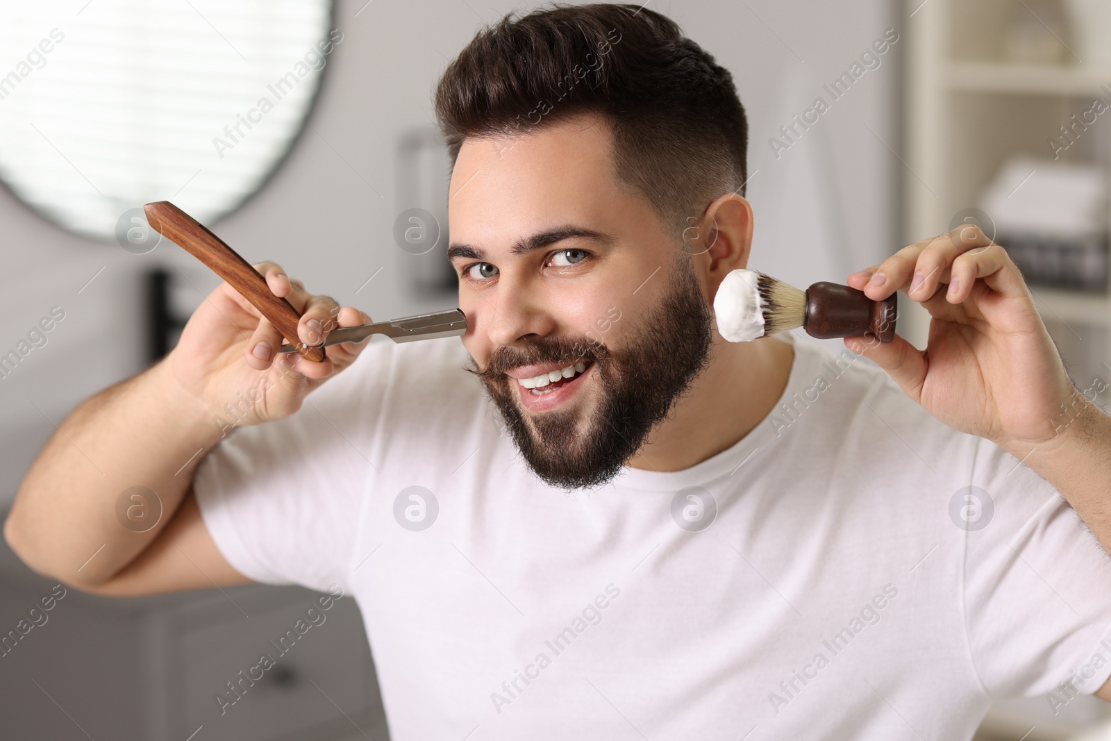 Photo of Handsome young man holding blade and shaving brush with foam in bathroom