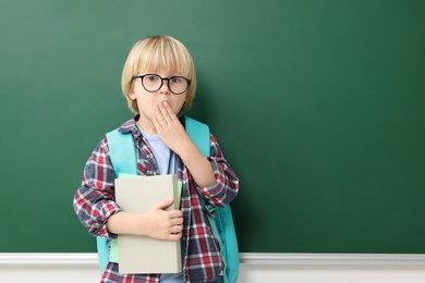 Photo of Emotional little school child with notebooks near chalkboard. Space for text