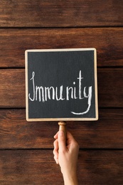 Photo of Woman holding small chalkboard with word Immunity against wooden background