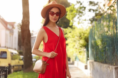 Beautiful young woman with stylish bag in red dress and sunglasses outdoors