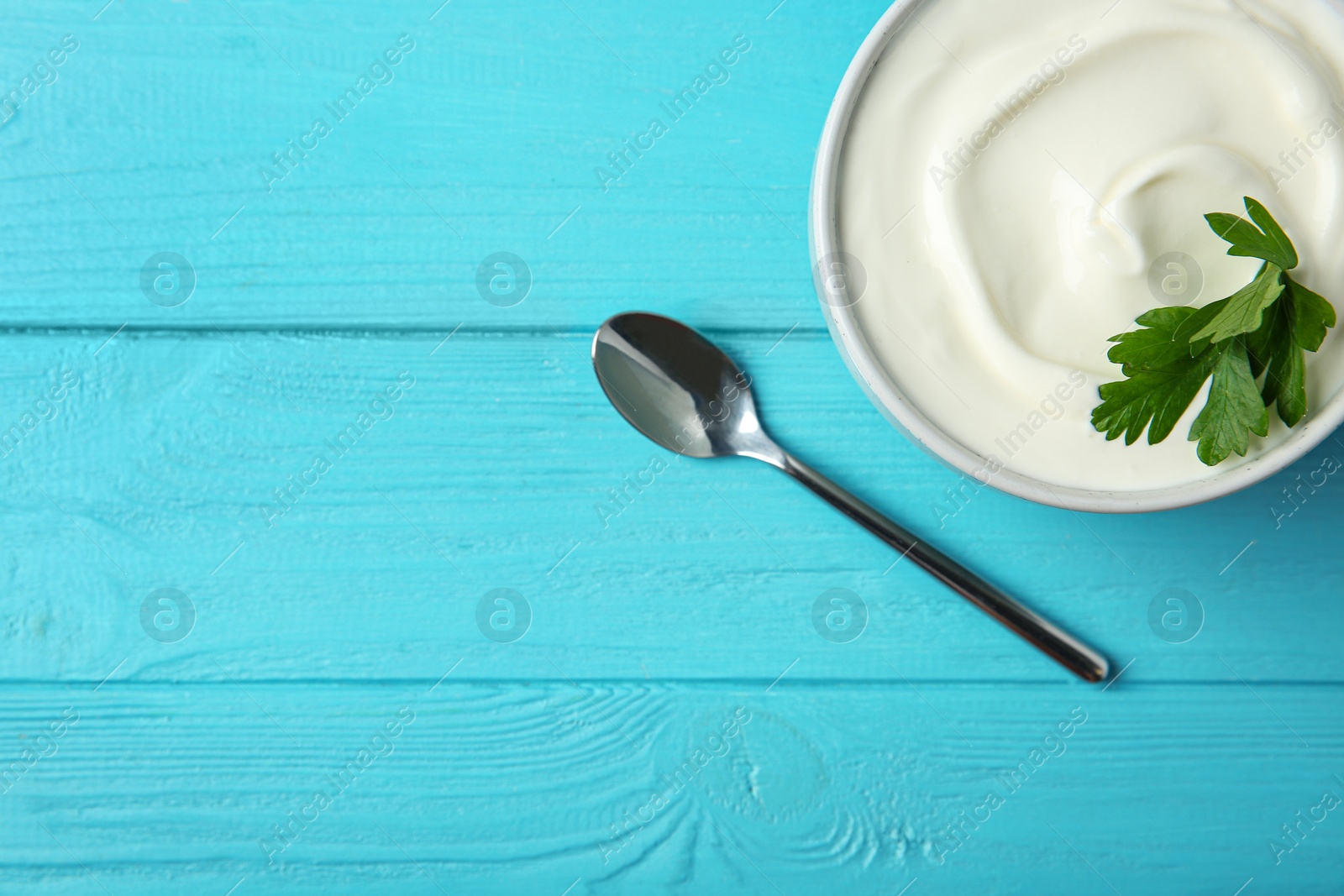 Photo of Bowl of fresh sour cream with parsley and spoon on light blue wooden table, flat lay. Space for text