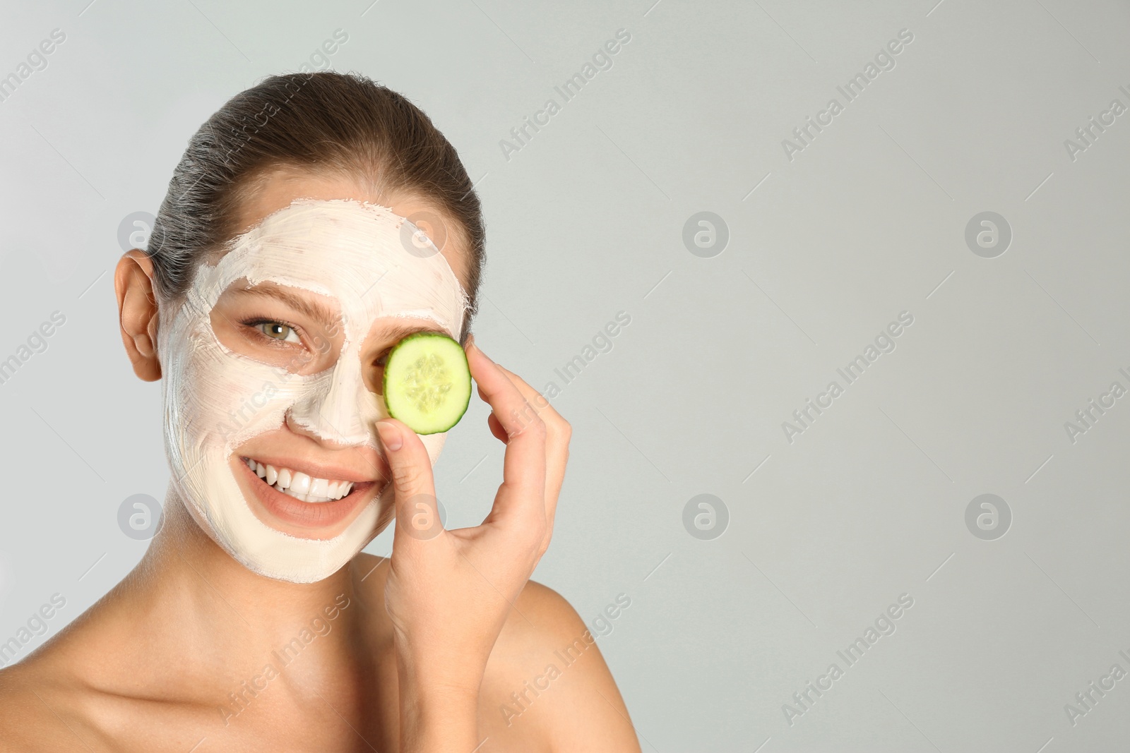 Photo of Beautiful woman holding cucumber slices near her face with clay mask against grey background. Space for text