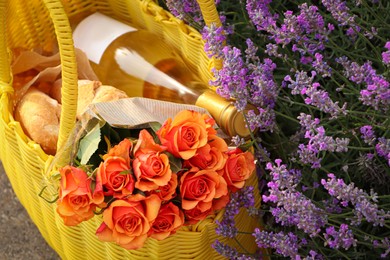 Yellow wicker bag with beautiful roses, bottle of wine and baguettes near lavender flowers outdoors, above view