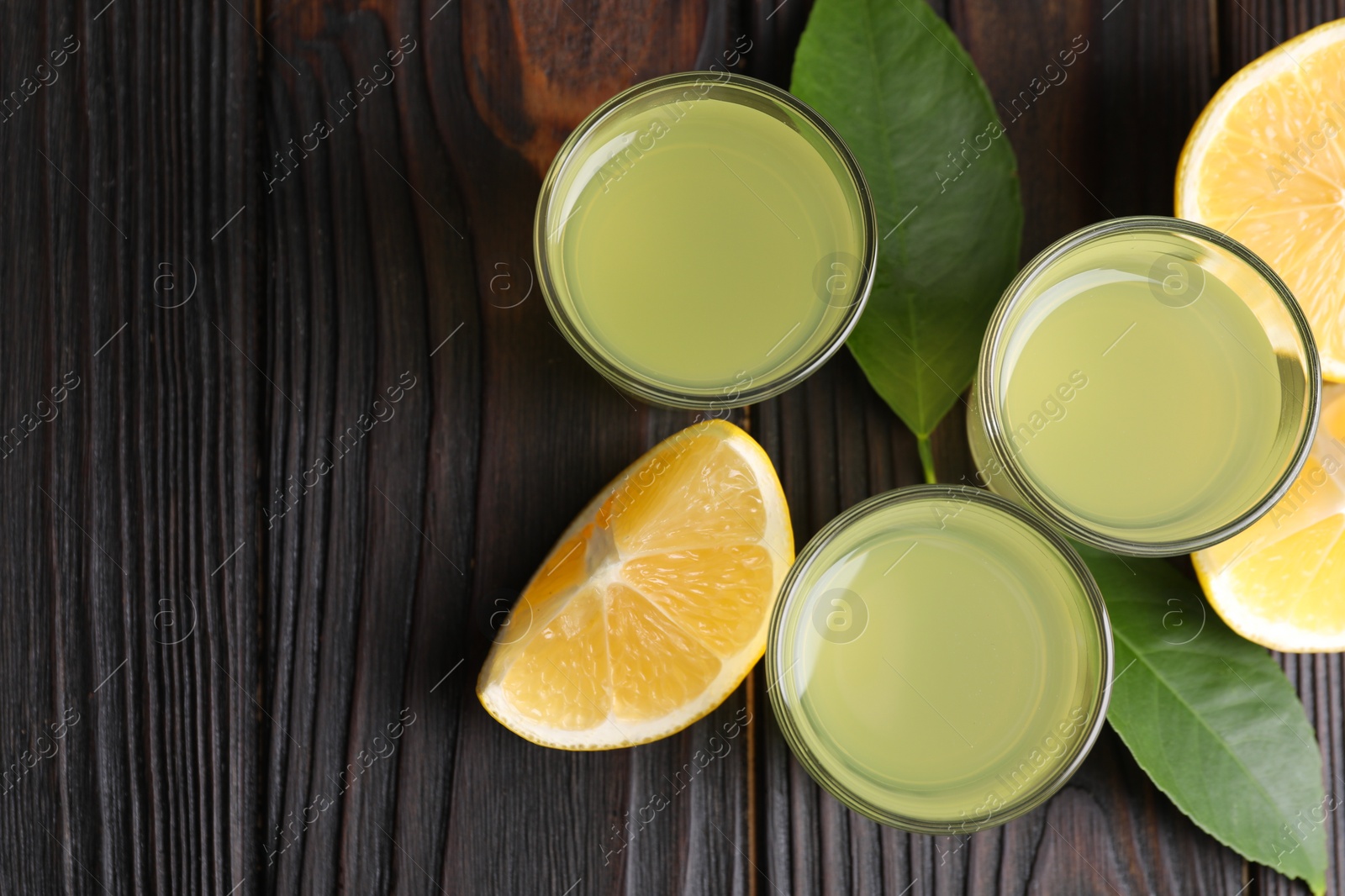 Photo of Tasty limoncello liqueur, lemon and green leaves on dark wooden table, flat lay. Space for text