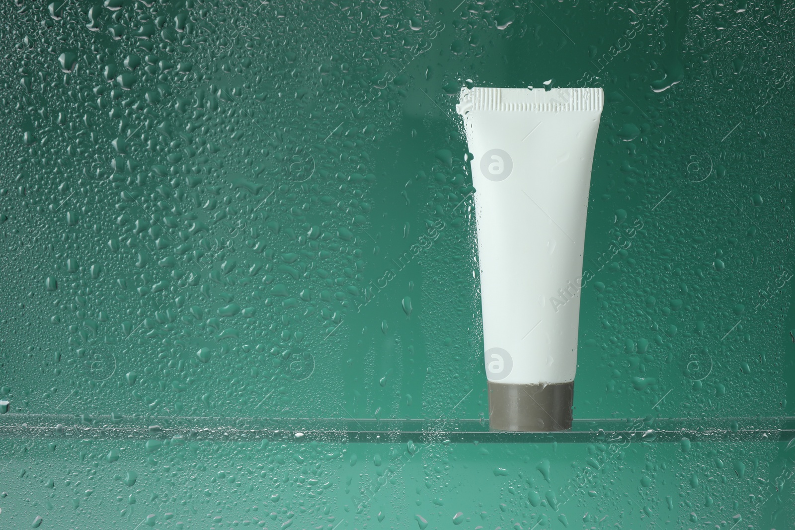 Photo of Tube with moisturizing cream on green background, view through wet glass. Space for text
