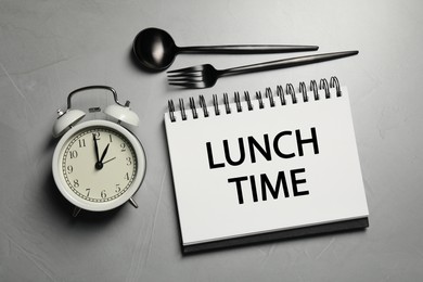 Image of Business lunch. Notebook with phrase Lunch Time, cutlery and alarm clock on light gray table, flat lay