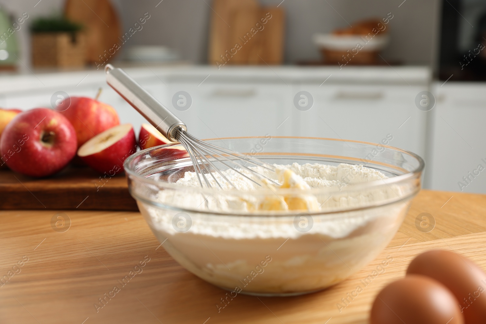 Photo of Cooking process. Metal whisk, bowl, flour, eggs and apple on wooden table in kitchen, closeup