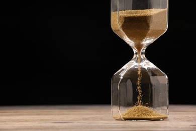 Hourglass with flowing sand on wooden table against black background, closeup. Space for text