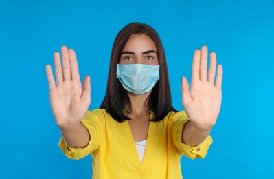Photo of Young woman in protective mask showing stop gesture on light blue background. Prevent spreading of coronavirus