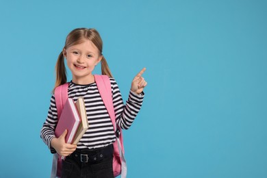 Photo of Happy schoolgirl with backpack and books pointing at something on light blue background, space for text