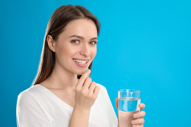 Photo of Young woman with glass of water taking vitamin pill on blue background